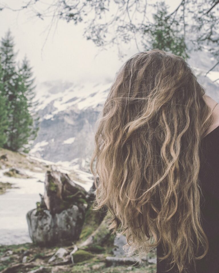 毛先が明るいブラウンの、くせを生かしたウェーブのロングヘア。雪の残る山を見る女性の後ろ姿